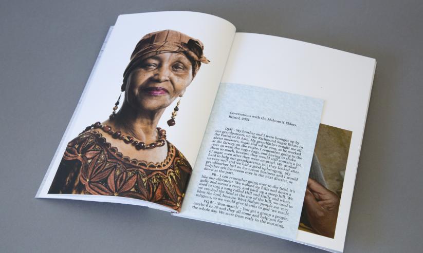 Image of the photobook, open on a page where there's a headshot of a stoic and friendly older Black women in traditional Jamaican clothes and head covering. She is smiling and has pink lipstick on with long earrings and a necklace. The tite of the pamphlet inside the book reads "Conversations with the Malcom X Elders. Bristol, 2021."