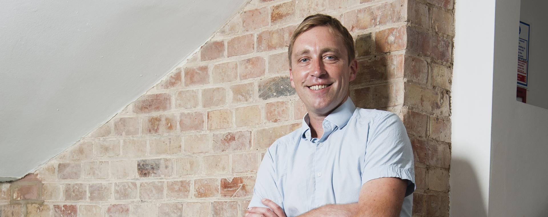 Conrad Peberdy standing infront of brick wall, arms folded, smiling