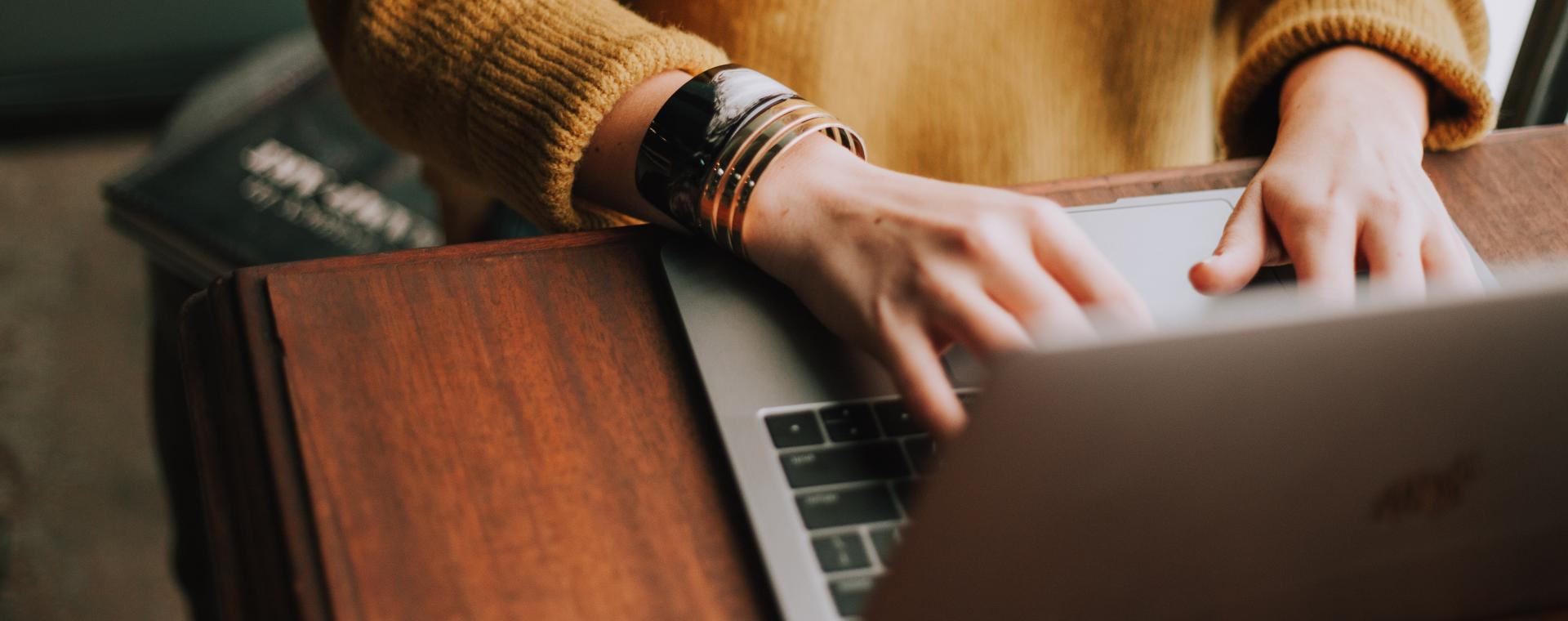 Person (face not visable) wearing a yellow jumper and a watch on their wrist working on a laptop