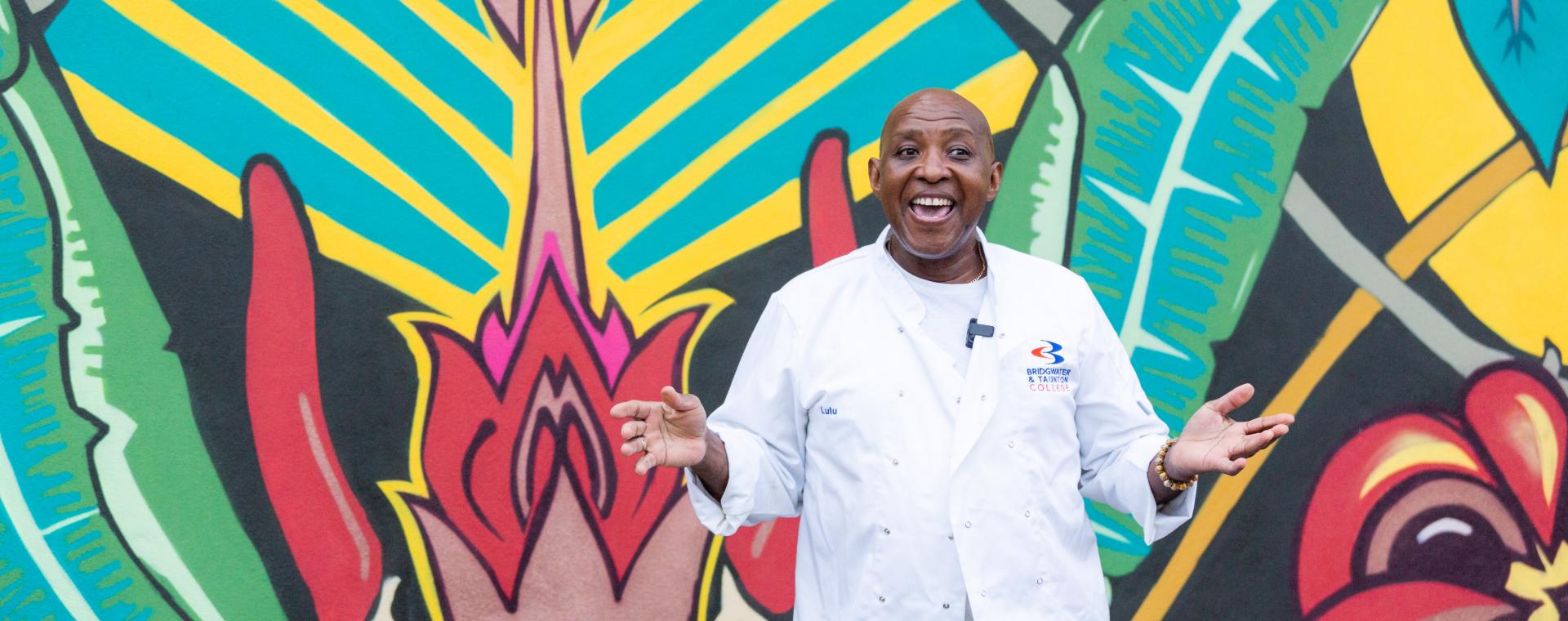 Glen from Glen's Kitchen standing in front of a brightly coloured garfitti mural, he is smiling with his hands out stretched palms up