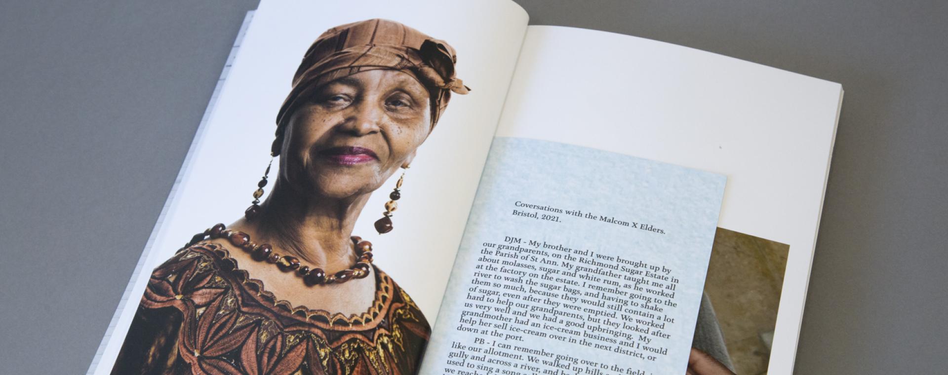 Image of the photobook, open on a page where there's a headshot of a stoic and friendly older Black women in traditional Jamaican clothes and head covering. She is smiling and has pink lipstick on with long earrings and a necklace. The tite of the pamphlet inside the book reads "Conversations with the Malcom X Elders. Bristol, 2021."