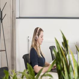 woman with blonde hair, with rose gold headpohones and grey t-shirt works on a laptop, in the foreround are green plants